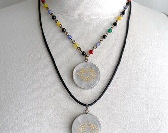 May include: Two-layered necklace with a spiritual theme. The top layer features a beaded chain with colorful beads. The bottom layer has a black cord with two round, clear pendants with a gold-colored geometric design.