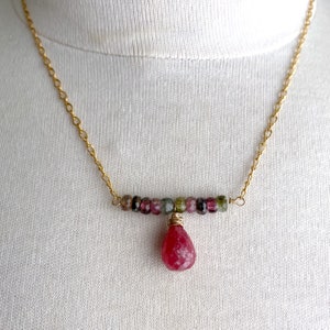 May include: A gold chain necklace with a pink and green gemstone pendant. The pendant is a teardrop shape and is made of multiple small gemstones.