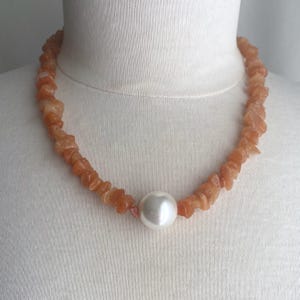May include: A necklace featuring a strand of irregular, tumbled orange stone chips arranged in a continuous loop, centered with a single, large, smooth, spherical white pearl that serves as the primary focal point of the jewelry piece.