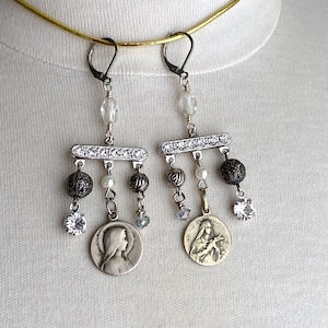May include: Silver dangle earrings with a religious charm, black beads, and clear crystals.