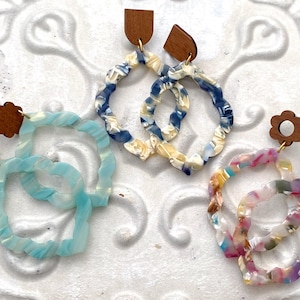 May include: Three pairs of earrings with unique designs. Each pair features a wooden top and a colorful, wavy, open-circle pendant. The earrings come in blue, multi-colored, and blue and white patterns, offering a variety of styles.