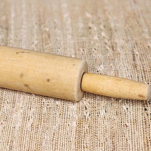 May include: A light-coloured wooden rolling pin with a cylindrical body and two handles. The rolling pin is made of natural wood and appears to be used for baking or cooking. The rolling pin is approximately 30 cm long.