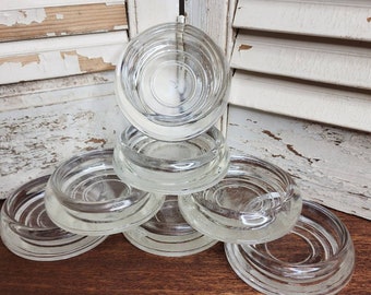 May include: A set of six clear glass coasters arranged in a stacked formation. Each coaster features a circular design with concentric rings pressed into the glass, creating a ridged texture on the surface and a raised outer rim for stability.