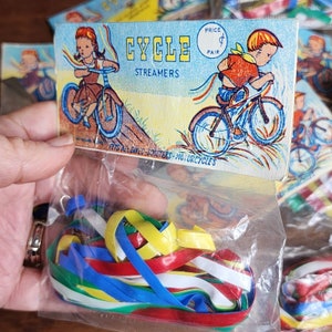 May include: Vintage bicycle streamers in a clear plastic bag. The packaging features illustrations of children riding bicycles and the words "CYCLE STREAMERS". The streamers are in red, yellow, blue, and green.