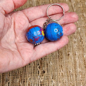 May include: A keychain with two small globe charms. The globes are blue with colourful continents and oceans. The keychain has a silver-coloured ring and chain. The globes are held in a person's hand.