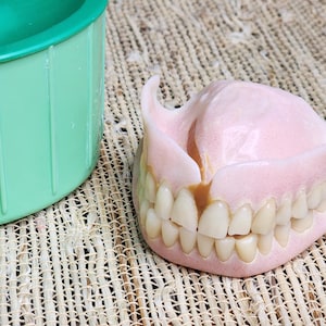 May include: A set of pink acrylic dentures with off-white teeth and a visible brown stain on one of the front teeth, positioned next to a green plastic container. The dentures feature a molded gum-colored base and a full arch of teeth.