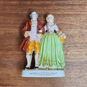 May include: A porcelain figurine of a couple standing side by side on a rectangular base. The figure on the left wears a brown coat, yellow breeches, and a white cravat. The figure on the right wears a pink bodice and a long, pleated green skirt.