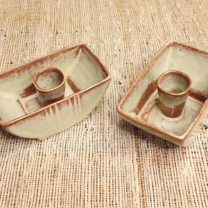 May include: Two handmade ceramic serving dishes with a light green glaze and brown trim. Each dish has a rectangular shape with a circular holder in the center. The dishes are placed on a textured, beige surface.