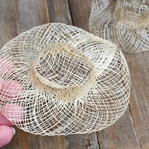 May include: Two small, circular, beige-colored hats crafted from a fine, open-weave straw material. Each hat features a flat, wide brim and a short, cylindrical crown with a hollow center, showcasing a delicate, grid-like texture throughout.