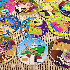 May include: A selection of circular souvenir coasters, each representing a different U.S. state. The coasters are adorned with state names, city names, and illustrations of landmarks and historical figures. They feature gold detailing and are displayed on a woven surface.
