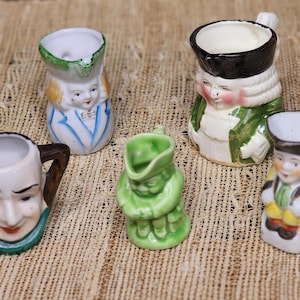 May include: A collection of five small, vintage ceramic pitchers. Each pitcher is uniquely designed with painted faces and clothing, featuring a variety of colors including white, green, yellow, and blue. The pitchers are arranged on a textured, tan surface.