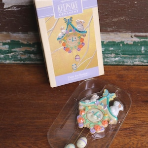 Könnte beinhalten: Hallmark Keepsake Osterschmuck in einer Schachtel. Das Ornament ist ein Kuckucksuhr-Design mit Hasen, Blumen und Ostereiern. Die Schachtel ist cremefarben mit einem lila Band und den Worten "Keepsake Ornament".