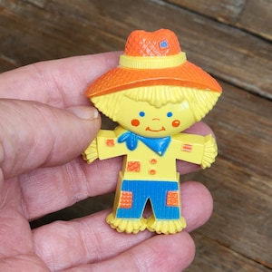 Puede incluir: Una figura de espantapájaros de plástico con un sombrero naranja brillante, cabello amarillo y una camisa amarilla con un pañuelo azul. La figura tiene pantalones azules con parches naranjas, mejillas rojas y ojos azules.