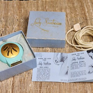 May include: Vintage Lady Sunbeam electric shaver in its original light blue box. The shaver is teal with a gold-colored decorative center. The box and instructions are included, along with the original power cord.