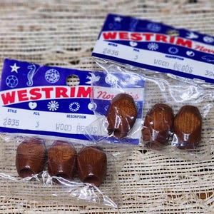 May include: Several packages of wood beads are displayed. Each package contains three dark brown, barrel-shaped beads. The packaging includes the text "WESTRIM Novelties" and "WOOD BEADS". The style number is 2835.