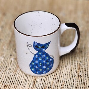 May include: White ceramic mug with a brown rim and handle. The mug features a blue cat design with white floral patterns. The handle is white with a brown accent. The mug has a speckled appearance.