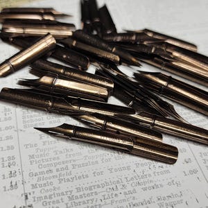 May include: A collection of vintage, bronze-colored pen nibs scattered on a printed paper background. The nibs vary in shape and size, with sharp, pointed tips. The paper has faded text, suggesting an antique or historical theme.