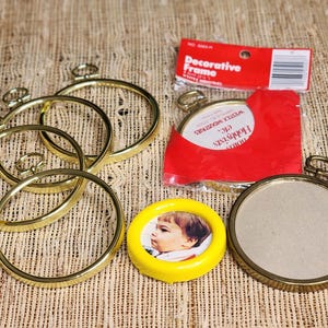 Könnte beinhalten: Eine Sammlung runder Rahmen, bestehend aus vier leeren, goldfarbenen Metallrahmen mit Aufhängeschlaufen, einem goldfarbenen Rahmen in einer roten Kunststoffverpackung mit der Aufschrift Decorative Frame und einem gelben Kunststoffrahmen mit Kinderfoto.
