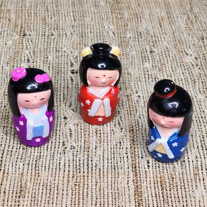 May include: Three small, decorative Japanese Kokeshi dolls. The dolls have black hair, painted faces, and wear colorful kimonos in purple, red, and blue. The dolls are on a textured, beige surface.