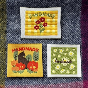 May include: Three embroidered patches with a yellow background and white stitching. The top patch has a yellow and white checkered pattern with red flowers and the text "HAND MADE". The middle patch has a black cat sitting in a field of orange flowers with green leaves and the text "HANDMADE". The bottom patch has a green background with white daisies and the text "handmade".