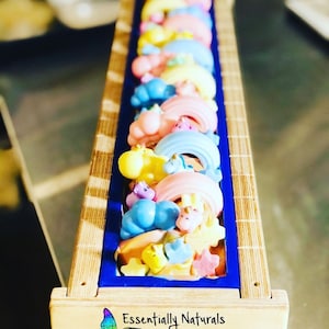 Può includere: Uno stampo in legno pieno di sapone colorato. Il sapone ha la forma di unicorni, arcobaleni e nuvole. Lo stampo è etichettato "Essentially Naturals Bath, Body & Skin Care".