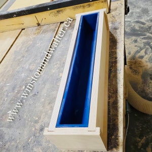 May include: A wooden mold for making soap, with a blue silicone liner. The mold is sitting on a wooden table. The text "www.winstonandwalter.ca" is visible on the table.