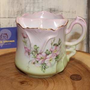 May include: A vintage porcelain shaving mug with a pink and green floral design. The mug has a gold rim and a handle with a floral design.