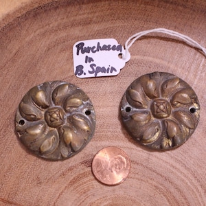 May include: Two antique brass round decorative accents with a floral design. The accents have a raised centre with a square design. The accents are approximately 5 cm in diameter. The text on the tag reads "Purchased in B. Spain".