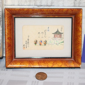 Puede incluir: Una obra de arte japonesa enmarcada que presenta una pequeña pagoda con un techo rojo y un grupo de personas con vestimenta tradicional. La obra de arte está sobre un fondo de color crema con caligrafía negra.