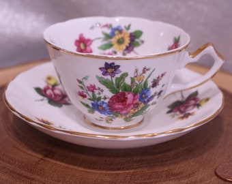 May include: A white teacup and saucer set with a floral design. The cup and saucer have a gold trim. The floral pattern includes pink, purple, blue, and yellow flowers. The teacup is approximately 3 inches in diameter.