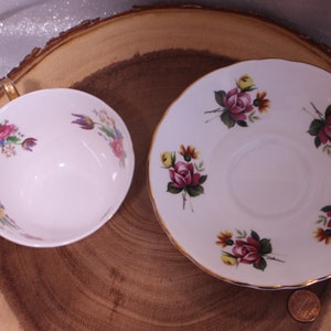 May include: A white teacup and saucer set with floral designs. The cup and saucer are decorated with pink, yellow, and purple flowers and green leaves. The cup has a gold handle and trim. The saucer has a gold rim.