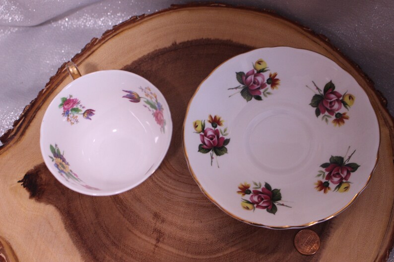 May include: A white teacup and saucer set with floral designs. The cup and saucer are decorated with pink, yellow, and purple flowers and green leaves. The cup has a gold handle and trim. The saucer has a gold rim.