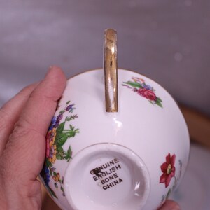 May include: White bone china teacup with a gold handle and floral pattern in red, pink, yellow, and blue. The bottom of the cup is stamped with "GENUINE ENGLISH BONE CHINA".