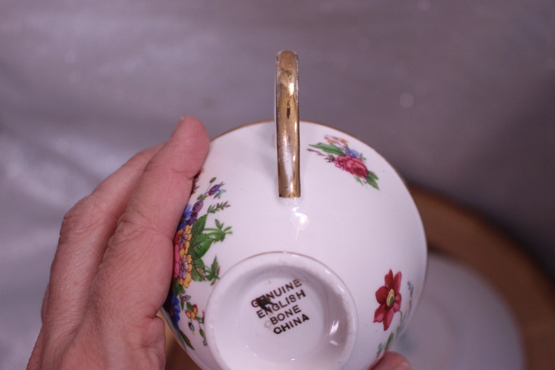 May include: White bone china teacup with a gold handle and floral pattern in red, pink, yellow, and blue. The bottom of the cup is stamped with "GENUINE ENGLISH BONE CHINA".