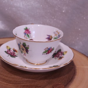 May include: White teacup and saucer set with floral patterns in pink, purple, and yellow. The edges of the cup and saucer are trimmed with gold. The teacup sits on a matching saucer, creating a classic tea set.