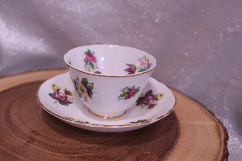May include: White teacup and saucer set with floral patterns in pink, purple, and yellow. The edges of the cup and saucer are trimmed with gold. The teacup sits on a matching saucer, creating a classic tea set.