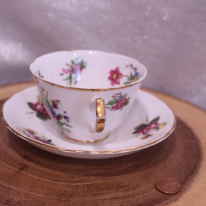 May include: A white teacup and saucer set with a floral pattern in pink, purple, and blue. The cup and saucer have gold trim. The set is displayed on a wooden surface.