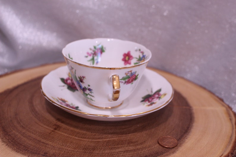 May include: A white teacup and saucer set with a floral pattern in pink, purple, and blue. The cup and saucer have gold trim. The set is displayed on a wooden surface.