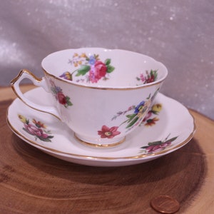 May include: White teacup and saucer set with a floral pattern in pink, red, blue, and yellow. The teacup and saucer have gold trim. The teacup is on a wooden surface.