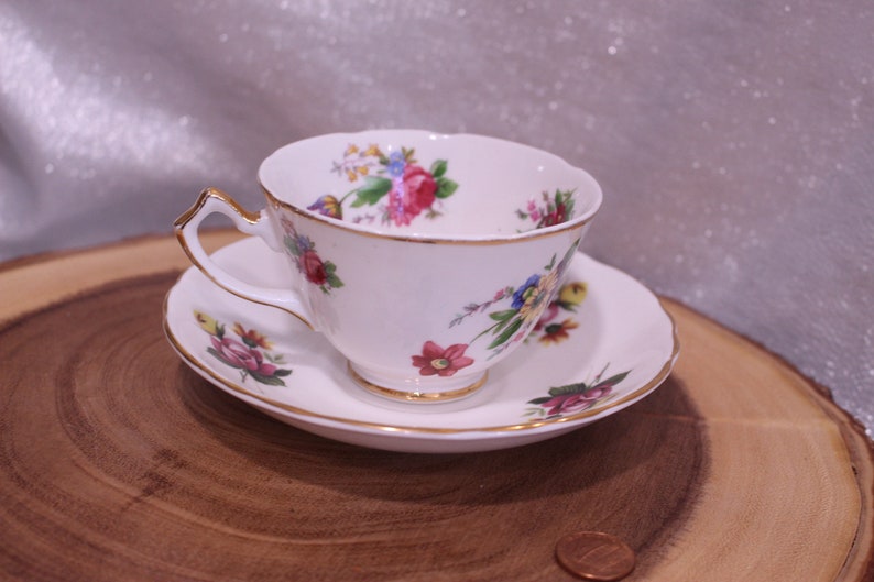 May include: White teacup and saucer set with a floral pattern in pink, red, blue, and yellow. The teacup and saucer have gold trim. The teacup is on a wooden surface.