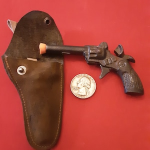 May include: A small, black metal toy gun with an orange tip and a brown leather holster. A silver quarter is included for scale.