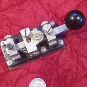 May include: Vintage telegraph key with a black knob and silver metal components. The key is mounted on a dark wooden base. A US quarter is included for size reference. The key is likely from the early to mid-20th century.