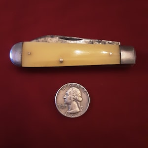 May include: A yellow handled pocket knife with two blades, closed, on a red surface. A silver US quarter coin is in the background.