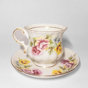 Op de afbeelding: Een witte theekop en schotel set met een bloemen rozenpatroon in roze, geel en paars. De kop en schotel hebben een gouden rand. De theekop heeft een gebogen handvat.