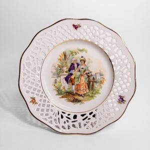 May include: A decorative porcelain plate with a scalloped edge and openwork design. The center features a painted scene of a couple in period clothing. The plate has a gold rim and is decorated with floral accents.