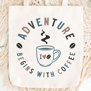 May include: Cream-colored tote bag with the text "ADVENTURE BEGINS WITH COFFEE" in a circular design. The design includes a coffee cup with a heart and coffee bean icons. The bag has cream-colored handles and the website address www.soverygraphic.com.