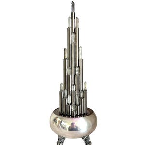 Könnte beinhalten: Eine silberfarbene dekorative Skulptur mit einer gestaffelten Anordnung von vertikalen Metallstäben und klaren kristallartigen Akzenten. Die Skulptur steht auf einem abgerundeten Sockel mit verzierten Füßen, ein eleganter Blickfang.
