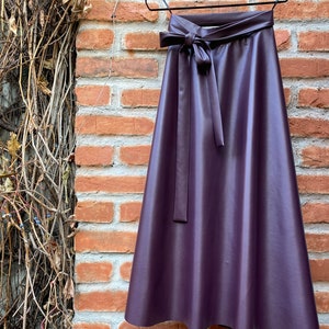 May include: A plum-colored, faux leather skirt hangs on a black hanger against a brick wall. The skirt features a high waist with a matching tie belt. The A-line skirt is a midi length.