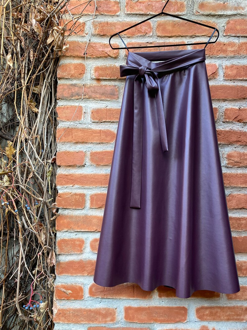 May include: A plum-colored, faux leather skirt hangs on a black hanger against a brick wall. The skirt features a high waist with a matching tie belt. The A-line skirt is a midi length.