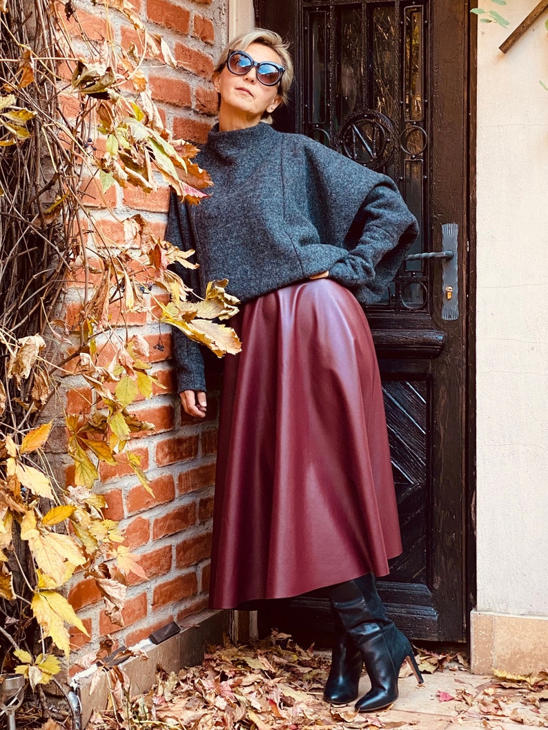 May include: A woman wearing a gray sweater, a burgundy leather skirt, and black boots. The skirt is knee-length and flared. The boots have a high heel. The background includes a brick wall and a dark wooden door.